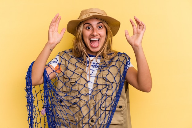Jonge kaukasische blonde vissersvrouw die een staaf houdt die op gele achtergrond wordt geïsoleerd en een aangename verrassing ontvangt, opgewonden en handen opsteekt.