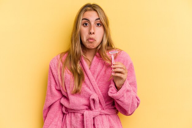 Jonge kaukasische blonde vrouw die een badjas draagt met bladen geïsoleerd op een gele achtergrond haalt haar schouders op en opent verwarde ogen.