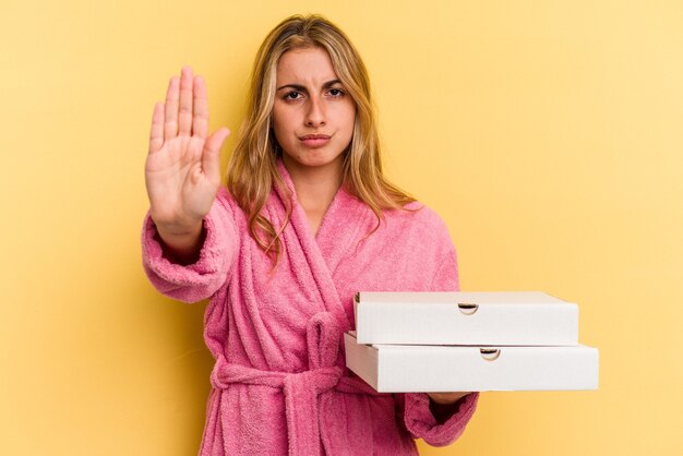 Jonge kaukasische blonde vrouw die een badjas draagt met pizza's geïsoleerd op een gele achtergrond die met uitgestrekte hand een stopbord toont, waardoor je wordt belet.