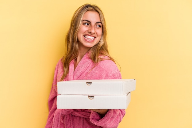 Jonge kaukasische blonde vrouw die een badjas draagt met pizza's geïsoleerd op een gele achtergrond ziet er glimlachend, vrolijk en aangenaam uit.