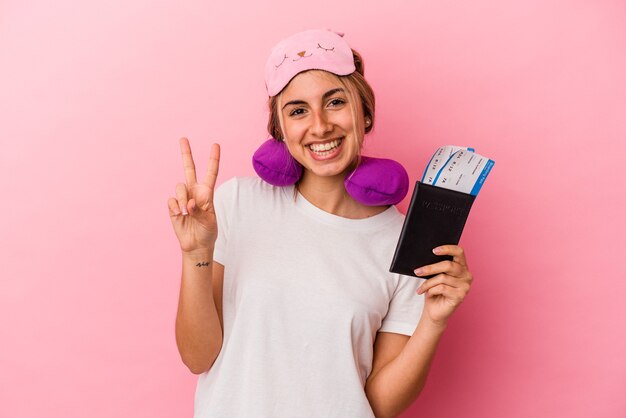 Jonge kaukasische blonde vrouw die een paspoort en kaartjes houdt om te reizen die op roze muur worden geïsoleerd die nummer twee met vingers toont