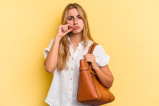 Jonge kaukasische blondevrouw die een leerzak draagt die op gele achtergrond met vingers op lippen wordt geïsoleerd die een geheim houden.