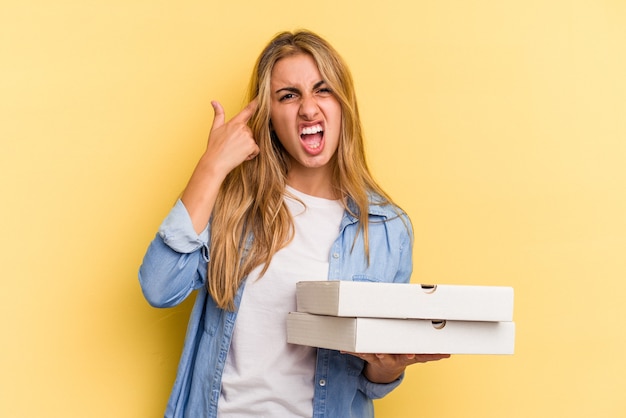 Jonge kaukasische blondevrouw die pizza's houdt die op gele achtergrond worden geïsoleerd die een teleurstellingsgebaar met wijsvinger tonen.