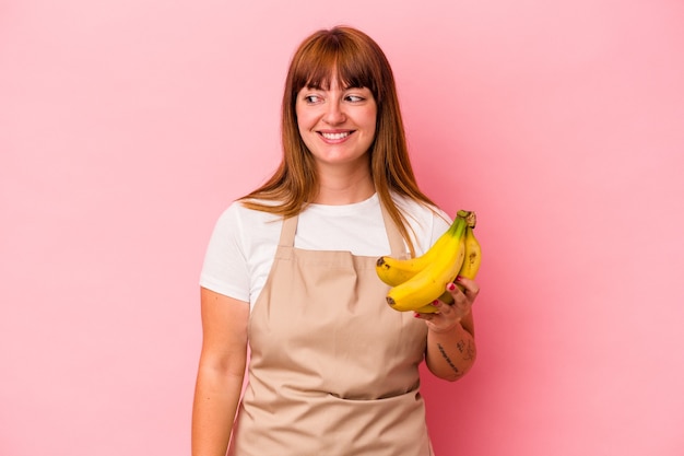 Jonge kaukasische bochtige vrouw die thuis kookt met bananen geïsoleerd op roze achtergrond ziet er glimlachend, vrolijk en aangenaam uit.