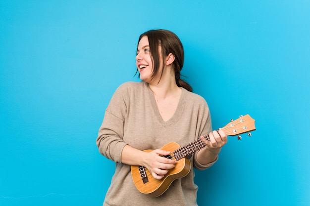 Jonge kaukasische bochtige vrouw die ukelele speelt die op een blauwe muur wordt geïsoleerd