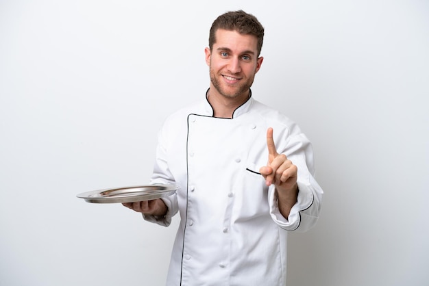 Jonge kaukasische chef-kok met dienblad dat op witte achtergrond wordt geïsoleerd die toont en een vinger opheft