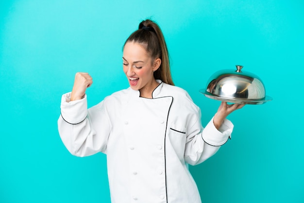 Jonge kaukasische chef-kokvrouw die met dienblad op blauwe achtergrond een overwinning viert