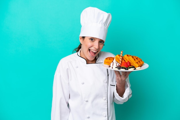 Jonge kaukasische chef-kokvrouw die wafels houdt die op blauwe achtergrond met verrassing en geschokte gezichtsuitdrukking worden geïsoleerd