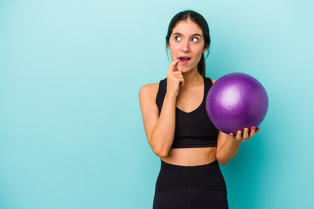 Jonge Kaukasische fitness vrouw met een bal geïsoleerd op blauwe achtergrond ontspannen denken over iets kijken naar een kopie ruimte.