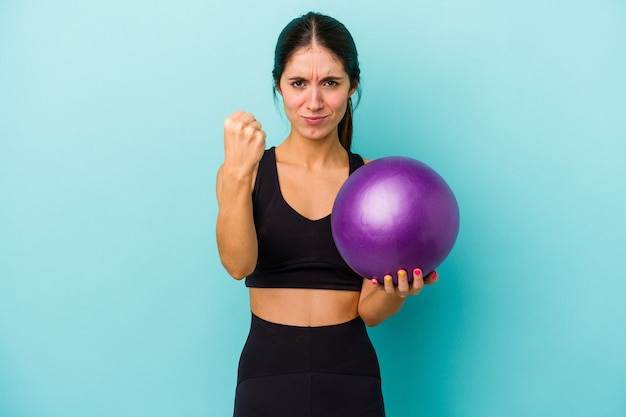 Jonge kaukasische fitnessvrouw die een bal houdt die op blauwe achtergrond wordt geïsoleerd die vuist toont aan camera, agressieve gezichtsuitdrukking.