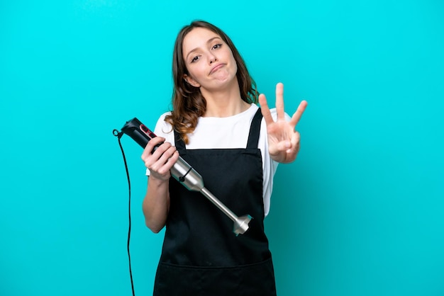 Jonge kaukasische fornuisvrouw die handblender gebruikt die op blauwe achtergrond gelukkig wordt geïsoleerd en drie met vingers telt