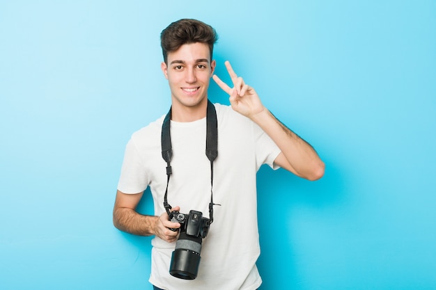 Jonge Kaukasische fotograafmens die een camera houdt die overwinningsteken toont en breed glimlacht.