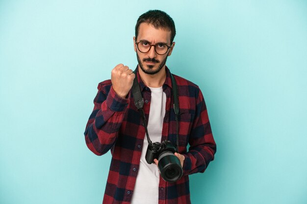 Jonge kaukasische fotograafmens die op blauwe achtergrond wordt geïsoleerd die vuist toont aan camera, agressieve gezichtsuitdrukking.