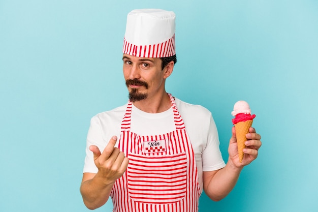 Jonge kaukasische ijsmaker die een ijsje vasthoudt dat op een blauwe achtergrond wordt geïsoleerd en met de vinger naar je wijst alsof uitnodigend dichterbij komt.