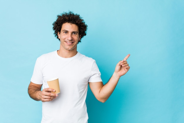 Jonge Kaukasische krullende mens die een meeneemkoffie houden glimlachend vrolijk wijzend met weg wijsvinger.