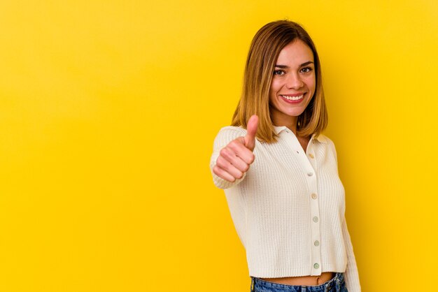 Jonge kaukasische magere vrouw die op gele muur wordt geïsoleerd die en duim opheft