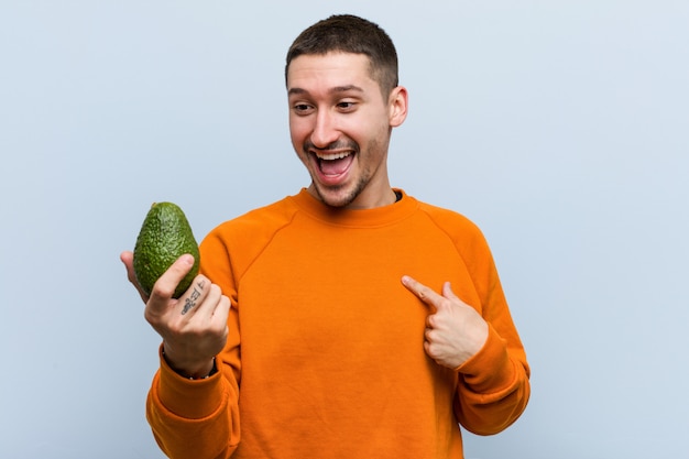 Jonge Kaukasische mens die een avocado houdt verraste richtend op zich, breed glimlachend.