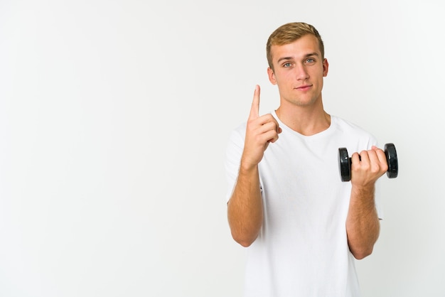 Jonge kaukasische mens die een gewicht houdt dat op witte achtergrond wordt geïsoleerd die nummer één met vinger toont.