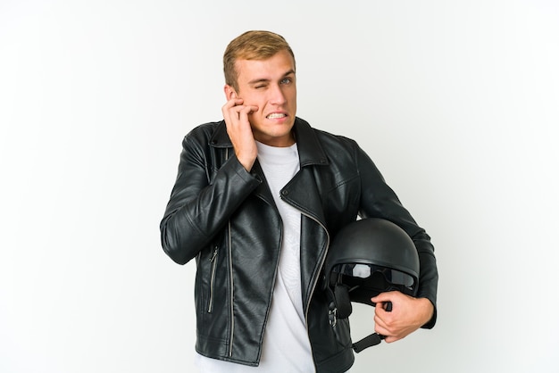 Jonge kaukasische mens die een motorhelm houdt die op witte achtergrond wordt geïsoleerd die oren behandelt met handen.