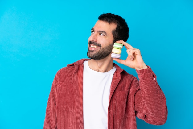 Jonge Kaukasische mens over geïsoleerde blauwe muur die kleurrijke Franse macarons houden en omhoog terwijl het glimlachen kijken