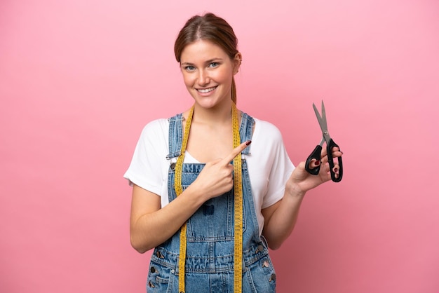 Jonge kaukasische naaistervrouw die op roze achtergrond wordt geïsoleerd die naar de kant wijst om een product te presenteren