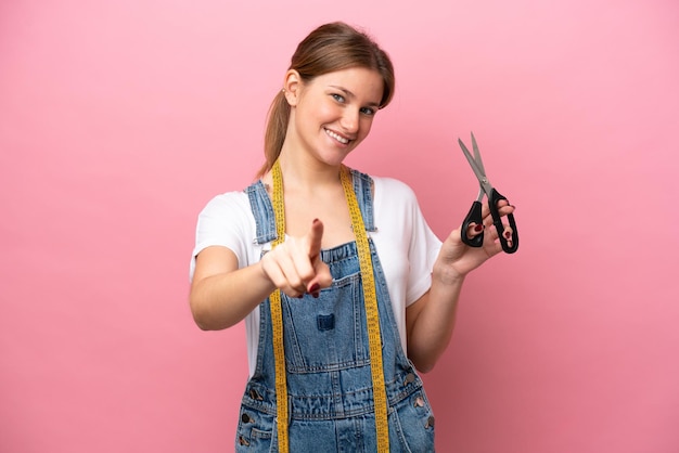 Jonge kaukasische naaistervrouw die op roze achtergrond wordt geïsoleerd die naar voren wijst met gelukkige uitdrukking