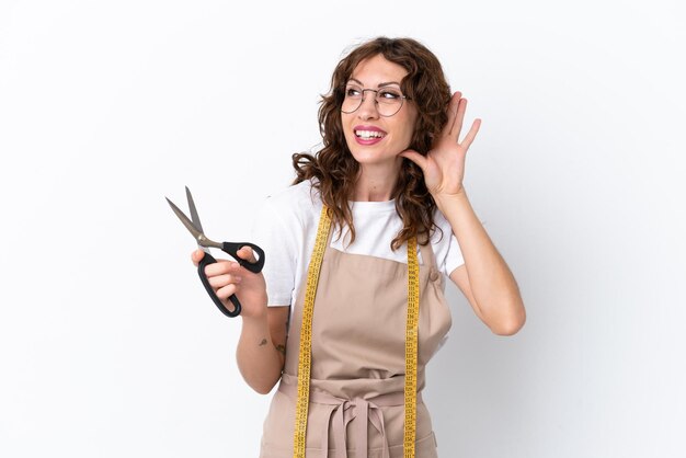 Jonge kaukasische naaistervrouw die op witte achtergrond wordt geïsoleerd die naar iets luistert door hand op het oor te leggen