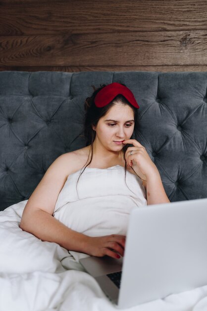 Jonge Kaukasische onderneemster die aan haar laptop in haar bed met nog slaapmasker werkt