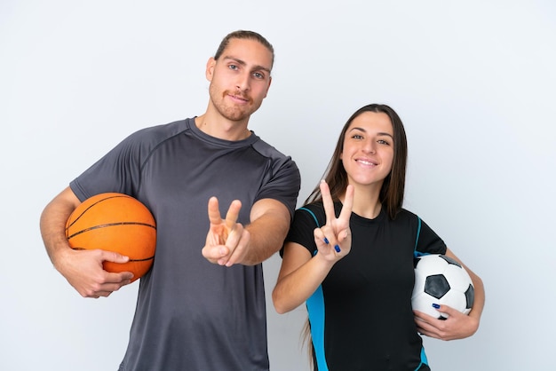 Jonge Kaukasische paar spelen basketbal en voetbal geïsoleerd op een witte achtergrond glimlachend en overwinning teken tonen
