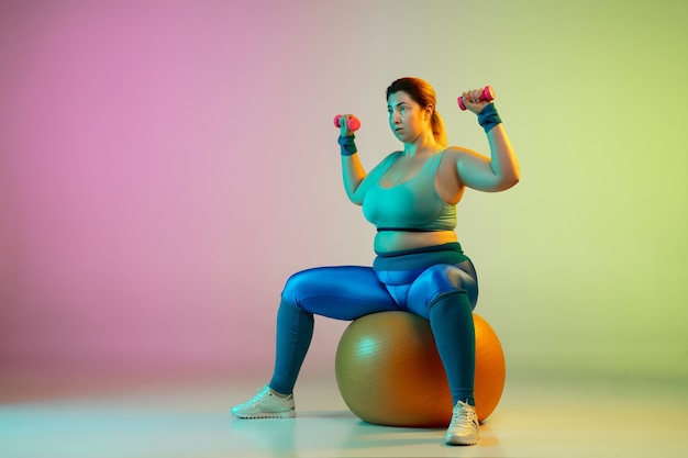 Jonge kaukasische plus size vrouwelijke model training op gradiënt paarse groene muur in neonlicht.