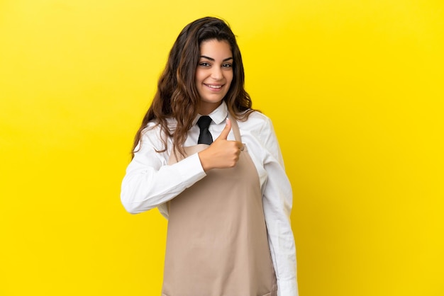 Jonge kaukasische restaurantkelner geïsoleerd op gele achtergrond die een duim omhoog gebaar geeft
