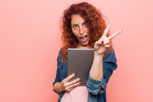 Jonge Kaukasische roodharigevrouw die een tablet houden die nummer twee met vingers tonen.