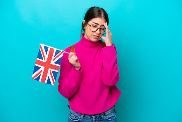Jonge kaukasische studentenvrouw die Engelse vlag houdt die op blauwe achtergrond met hoofdpijn wordt geïsoleerd