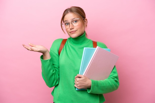 Jonge kaukasische studentenvrouw die op roze achtergrond wordt geïsoleerd die twijfels heeft terwijl het opheffen van handen