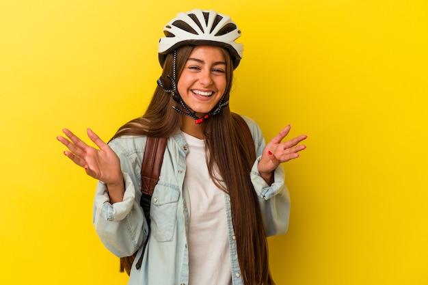 Jonge kaukasische studentvrouw die een fietshelm draagt die op gele achtergrond wordt geïsoleerd en een aangename verrassing ontvangt, opgewonden en handen opsteekt.