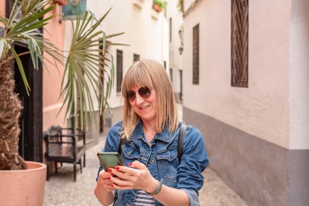 Jonge kaukasische toeristenvrouw in spijkerjasje die lacht met smartphone op straat in de stad