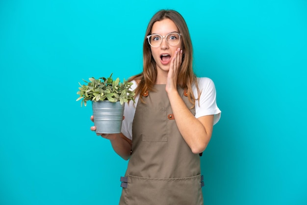 Jonge kaukasische tuinmanvrouw die een installatie houdt die op blauwe achtergrond met verrassing en geschokte gezichtsuitdrukking wordt geïsoleerd