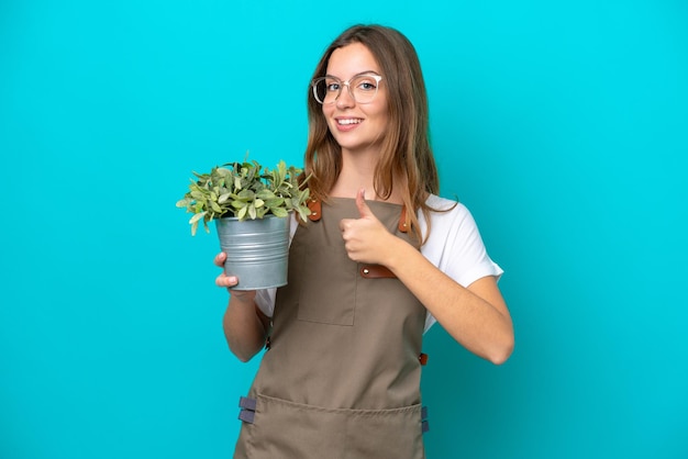 Jonge kaukasische tuinmanvrouw die een installatie houdt die op blauwe achtergrond wordt geïsoleerd en een duim omhoog gebaar geeft