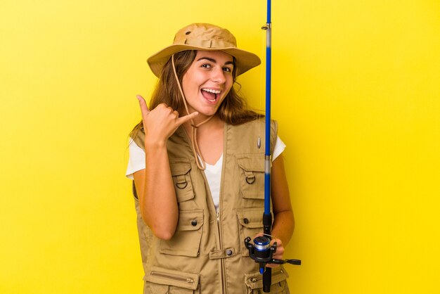 Jonge kaukasische vissersvrouw die een staaf houdt die op gele achtergrond wordt geïsoleerd die een mobiel telefoongesprekgebaar met vingers toont.