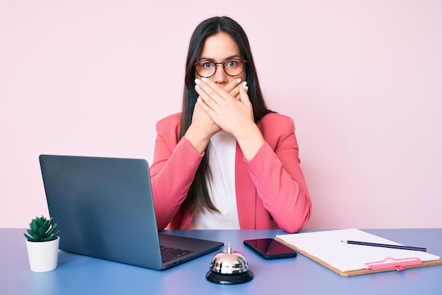 Jonge kaukasische vrouw die aan het bureau van de recepcionist zit en met behulp van een laptop werkt, bedek de mond met de handen voor een fout geheim concept