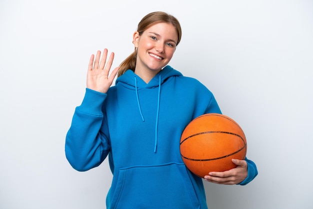 Jonge kaukasische vrouw die basketbal speelt geïsoleerd op een witte achtergrond die met de hand salueert met een gelukkige uitdrukking