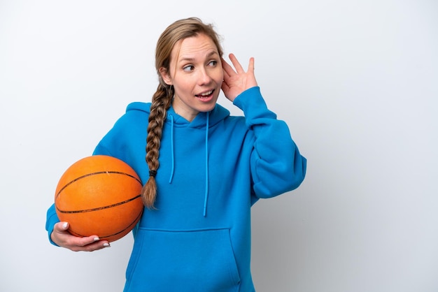 Jonge kaukasische vrouw die basketbal speelt geïsoleerd op een witte achtergrond die naar iets luistert door de hand op het oor te leggen