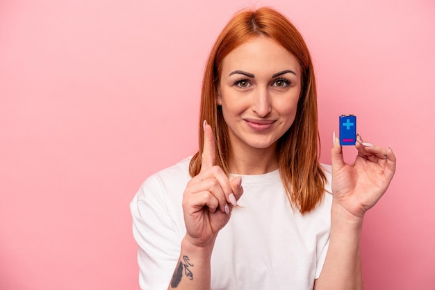 Jonge kaukasische vrouw die batterijen houdt die op roze achtergrond wordt geïsoleerd die nummer één met vinger toont.