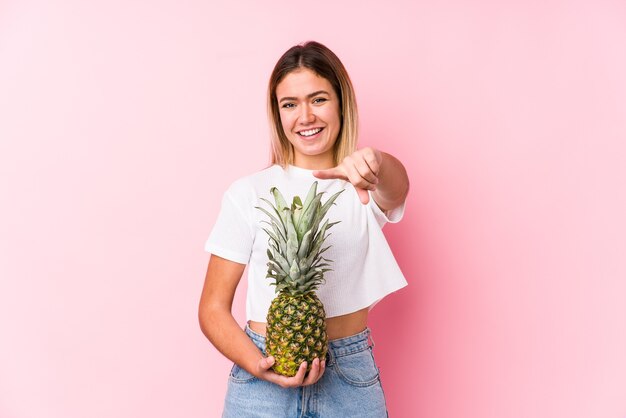 Jonge kaukasische vrouw die een ananas vrolijke glimlach houdt die naar voorzijde wijst.