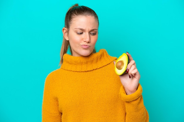 Jonge kaukasische vrouw die een avocado houdt die op blauwe achtergrond met droevige uitdrukking wordt geïsoleerd