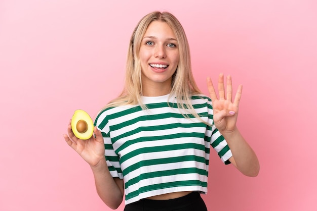 Jonge kaukasische vrouw die een avocado houdt die op roze achtergrond gelukkig wordt geïsoleerd en vier met vingers telt