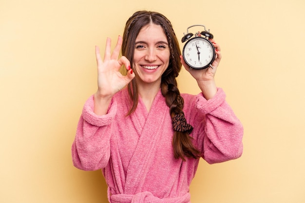 Jonge kaukasische vrouw die een badjas draagt die een wekker houdt die op gele achtergrond wordt geïsoleerd, vrolijk en zelfverzekerd toont een goed gebaar.