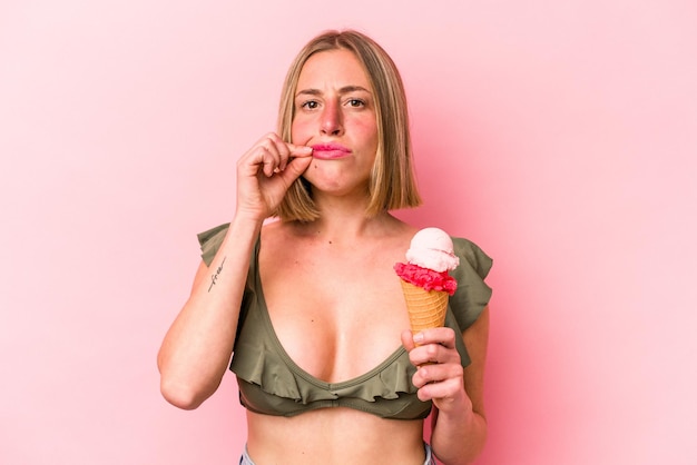 Jonge kaukasische vrouw die een bikini draagt en een roomijs houdt dat op roze achtergrond wordt geïsoleerd met vingers op lippen die een geheim houden