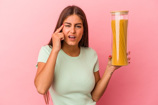 Jonge kaukasische vrouw die een deegwarenkruik houdt die op roze achtergrond wordt geïsoleerd die een teleurstellingsgebaar met wijsvinger toont.