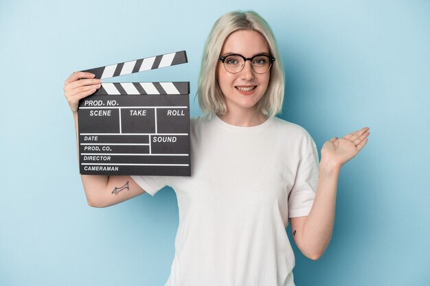 Jonge kaukasische vrouw die een filmklapper houdt die op blauwe achtergrond wordt geïsoleerd die een exemplaarruimte op een palm toont en een andere hand op taille houdt.